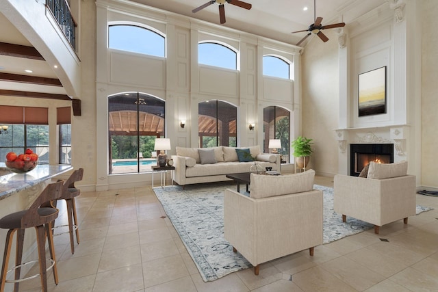 living room with a towering ceiling, ceiling fan, light tile patterned floors, a premium fireplace, and beamed ceiling