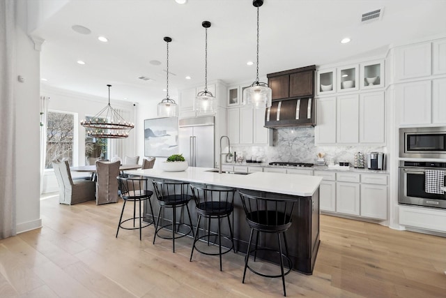 kitchen with sink, pendant lighting, built in appliances, light hardwood / wood-style floors, and an island with sink