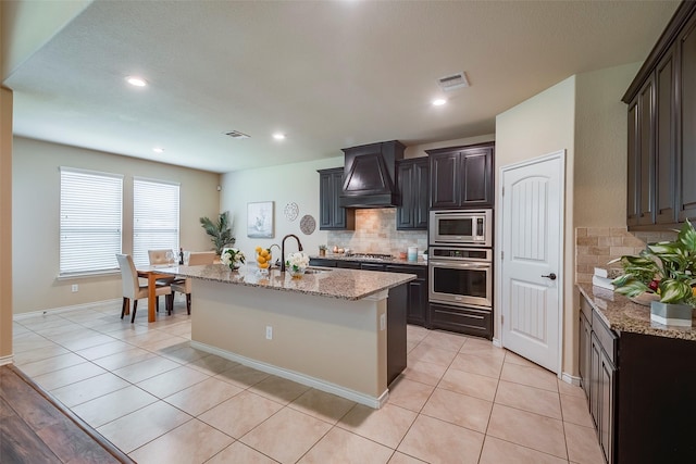 kitchen with appliances with stainless steel finishes, an island with sink, sink, custom exhaust hood, and light stone countertops