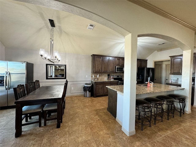 kitchen with light stone counters, hanging light fixtures, dark brown cabinets, appliances with stainless steel finishes, and an island with sink