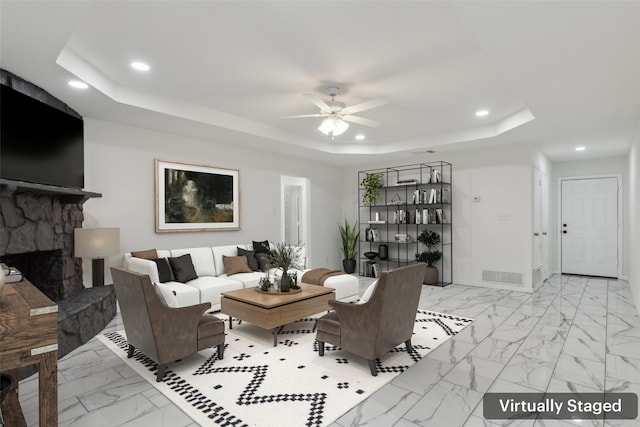 living room with a raised ceiling, a stone fireplace, and ceiling fan