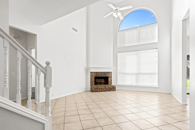 unfurnished living room with a brick fireplace, ceiling fan, light tile patterned floors, a high ceiling, and plenty of natural light