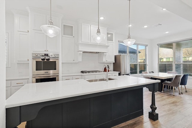 kitchen featuring backsplash, hanging light fixtures, stainless steel appliances, white cabinets, and a center island with sink