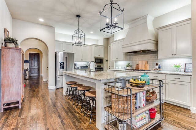 kitchen with appliances with stainless steel finishes, backsplash, custom exhaust hood, pendant lighting, and a center island with sink