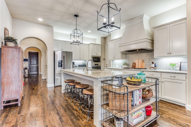 kitchen featuring dark wood-type flooring, a center island with sink, custom range hood, pendant lighting, and stainless steel appliances