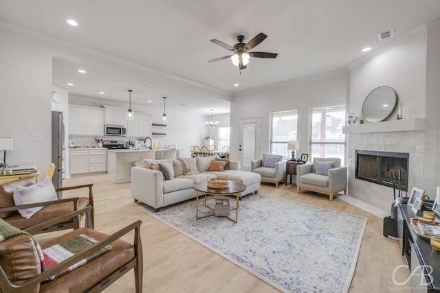 living room with light wood-type flooring, ornamental molding, ceiling fan with notable chandelier, sink, and a tiled fireplace