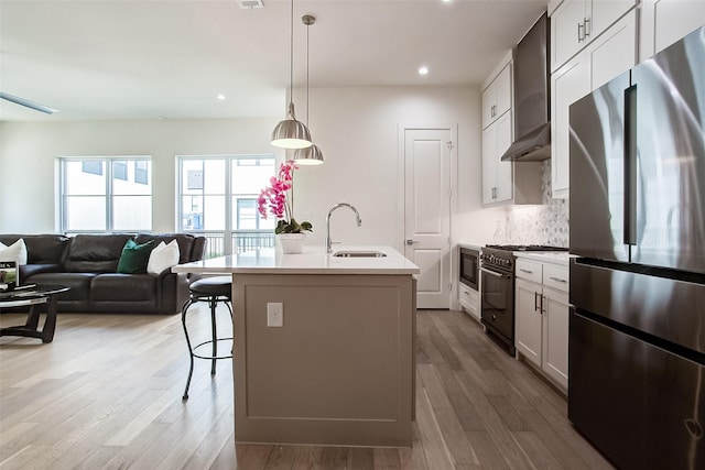 kitchen with wall chimney exhaust hood, sink, white cabinetry, appliances with stainless steel finishes, and a kitchen island with sink