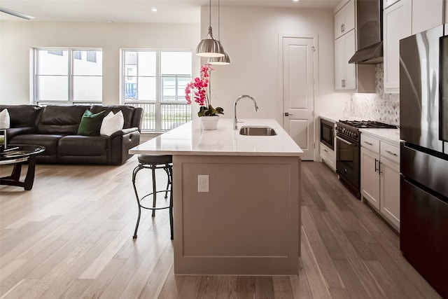 kitchen with sink, hanging light fixtures, stainless steel appliances, a center island with sink, and wall chimney exhaust hood