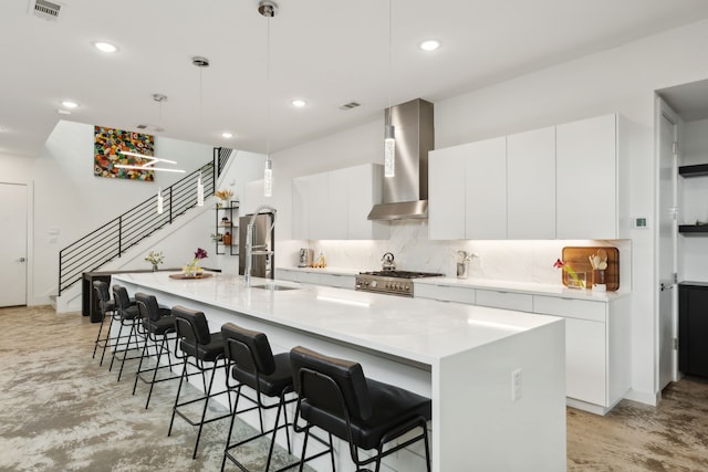 kitchen featuring decorative light fixtures, an island with sink, wall chimney range hood, decorative backsplash, and white cabinets