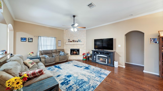 living room with dark wood-type flooring, a stone fireplace, ceiling fan, and crown molding