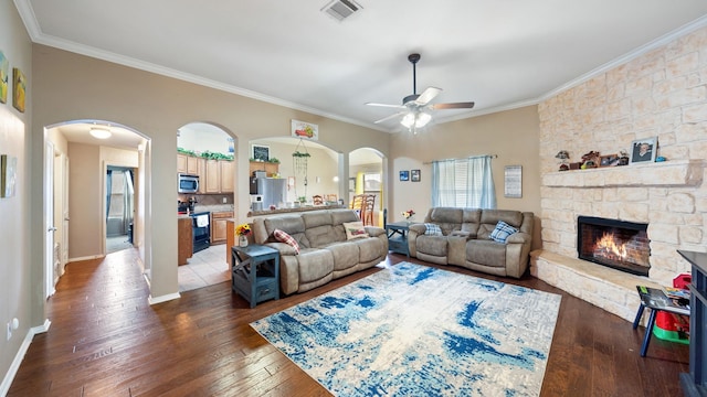 living room featuring ceiling fan, a stone fireplace, crown molding, and dark wood-type flooring