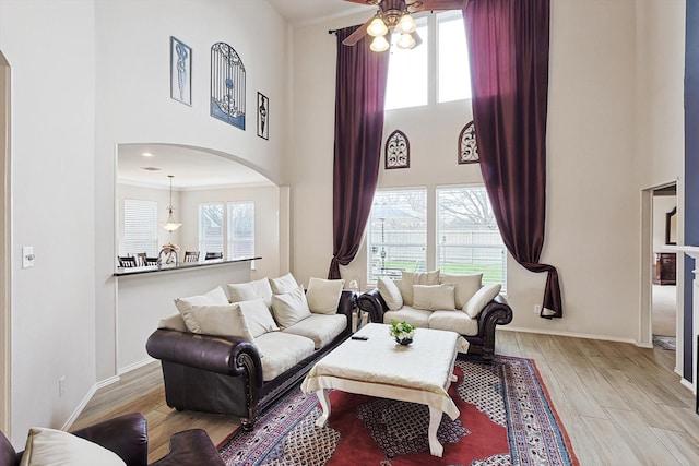 living room with baseboards, light wood-style flooring, arched walkways, ceiling fan, and a towering ceiling