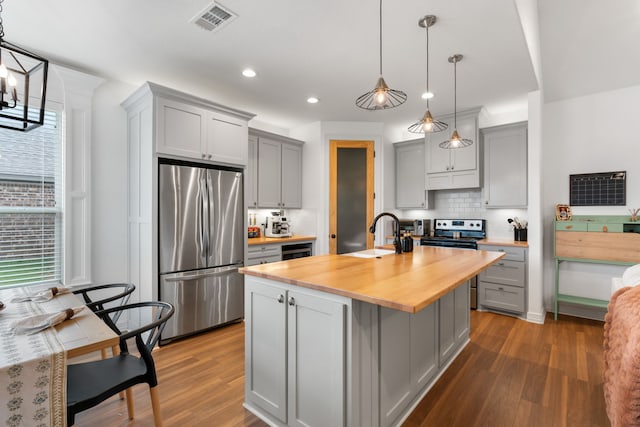 kitchen featuring gray cabinetry, a kitchen island with sink, hanging light fixtures, appliances with stainless steel finishes, and butcher block countertops