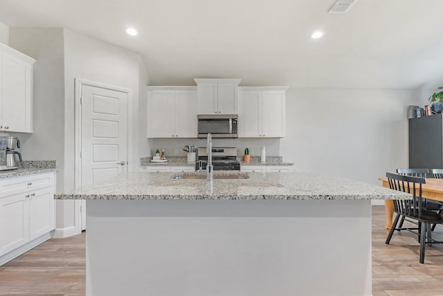 kitchen featuring stainless steel appliances, a kitchen island with sink, light wood-type flooring, white cabinets, and light stone counters