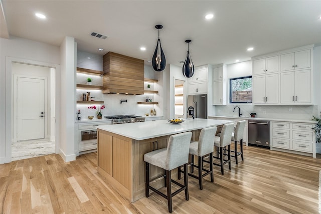 kitchen with premium range hood, a center island with sink, appliances with stainless steel finishes, light hardwood / wood-style floors, and white cabinetry