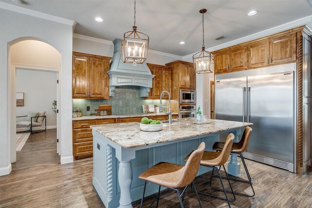 kitchen with custom exhaust hood, a center island with sink, built in appliances, tasteful backsplash, and decorative light fixtures