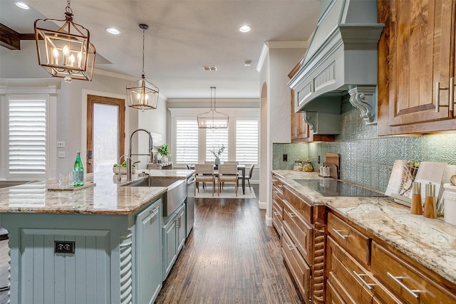 kitchen with black electric stovetop, light stone counters, a kitchen island with sink, crown molding, and sink