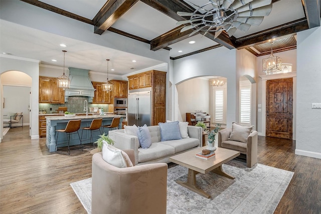 living room featuring light wood-type flooring, ceiling fan with notable chandelier, crown molding, sink, and beamed ceiling