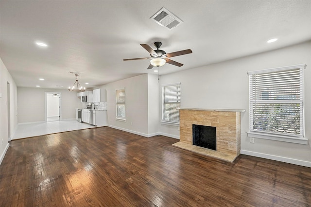 unfurnished living room with a stone fireplace, a wealth of natural light, dark hardwood / wood-style floors, and ceiling fan with notable chandelier