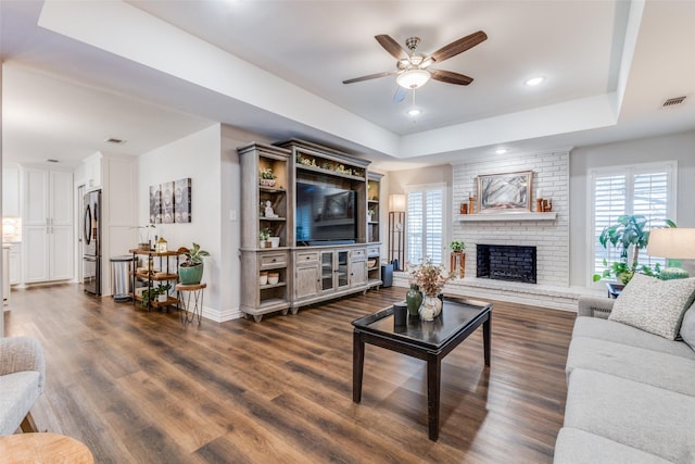 living room with ceiling fan, a tray ceiling, dark hardwood / wood-style flooring, and a brick fireplace