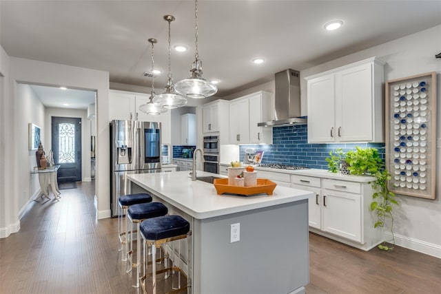 kitchen with an island with sink, appliances with stainless steel finishes, decorative light fixtures, wall chimney exhaust hood, and white cabinets