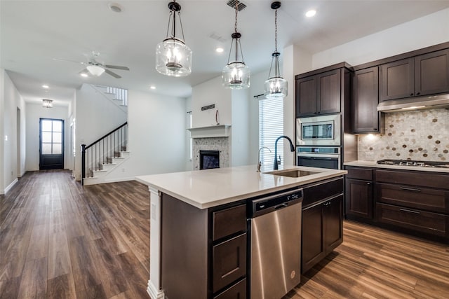 kitchen with appliances with stainless steel finishes, backsplash, a kitchen island with sink, dark brown cabinets, and sink