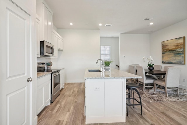 kitchen with sink, appliances with stainless steel finishes, a kitchen island with sink, light stone countertops, and white cabinets