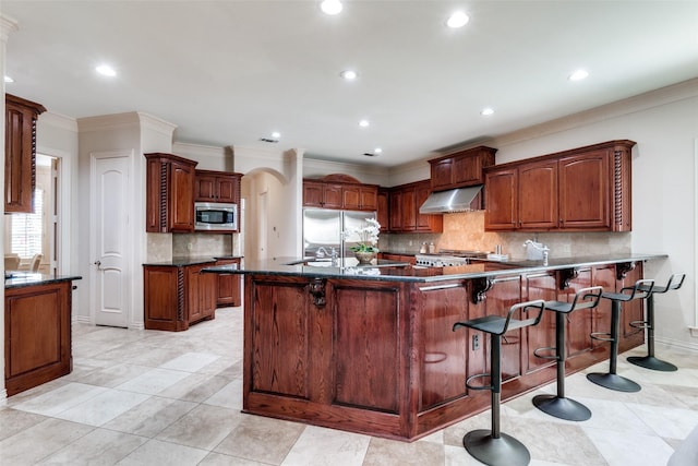 kitchen with a kitchen breakfast bar, ornamental molding, exhaust hood, built in appliances, and kitchen peninsula