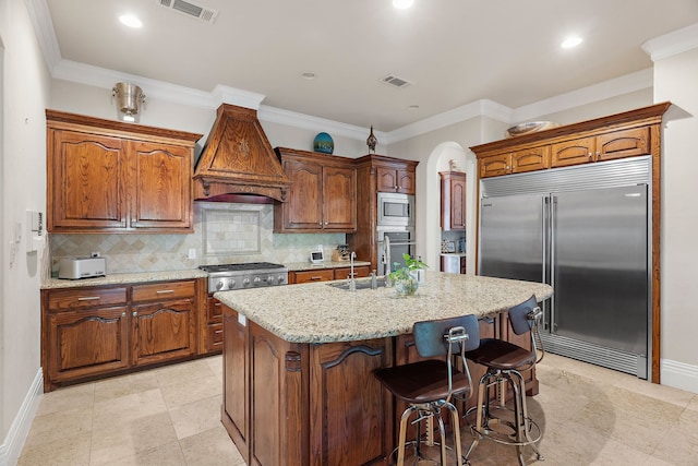 kitchen featuring backsplash, ornamental molding, custom exhaust hood, a kitchen island with sink, and built in appliances