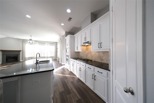 kitchen with a fireplace, a sink, white cabinets, under cabinet range hood, and dark countertops