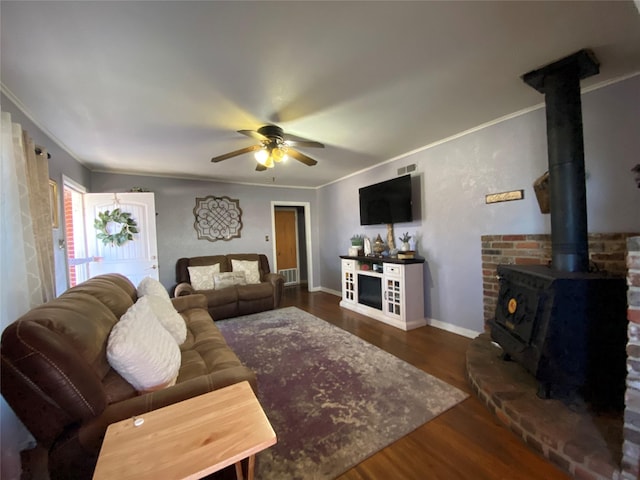 living area featuring ceiling fan, baseboards, ornamental molding, dark wood-style floors, and a wood stove