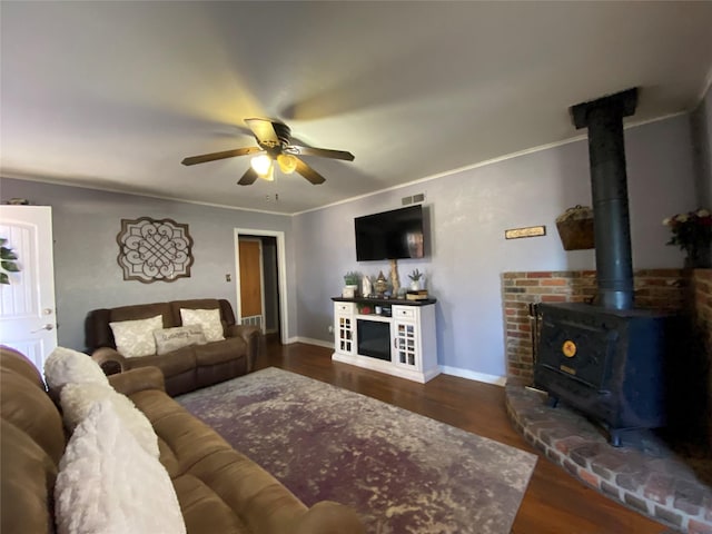 living room with dark wood-style flooring, crown molding, a ceiling fan, a wood stove, and baseboards