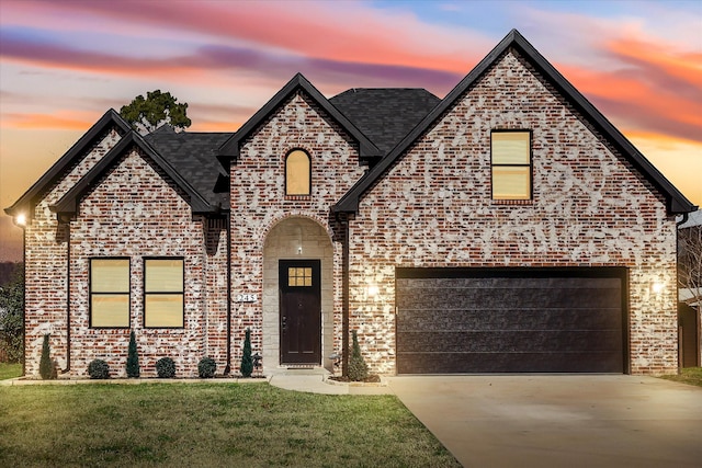 french country home featuring an attached garage, a lawn, a shingled roof, brick siding, and driveway