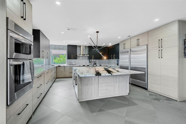 kitchen featuring a center island with sink, wall chimney exhaust hood, light stone countertops, decorative light fixtures, and stainless steel appliances