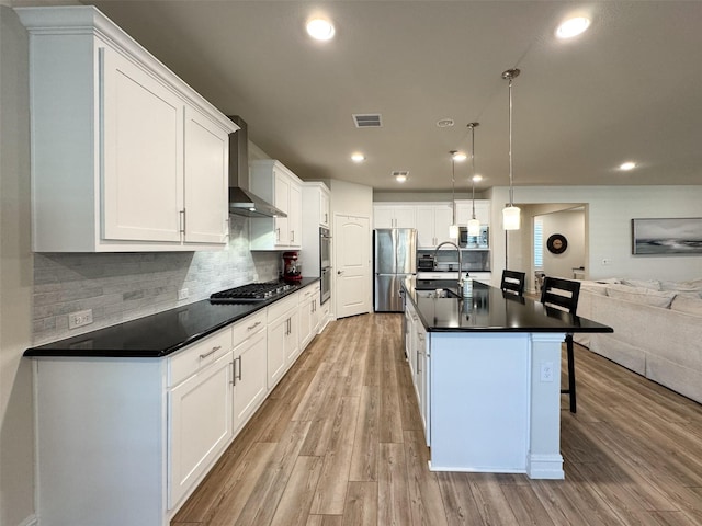 kitchen with stainless steel appliances, wall chimney range hood, pendant lighting, white cabinets, and an island with sink