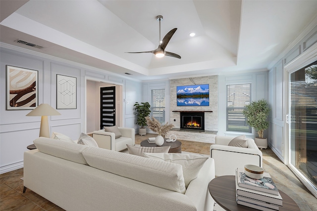 living room featuring ornamental molding, a stone fireplace, ceiling fan, and a tray ceiling