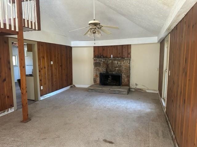 unfurnished living room featuring ceiling fan, a stone fireplace, wood walls, carpet floors, and a textured ceiling