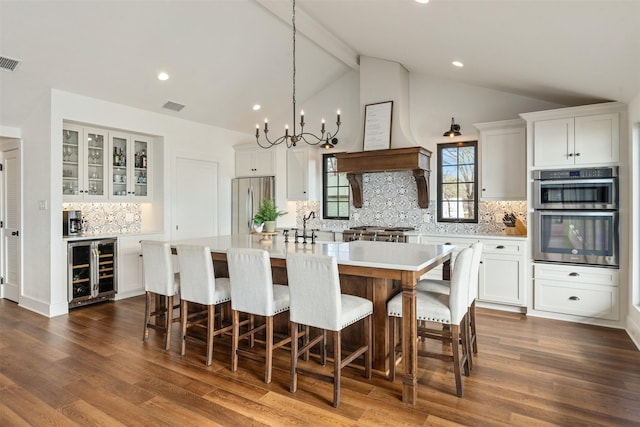 kitchen featuring white cabinetry, beverage cooler, stainless steel appliances, vaulted ceiling with beams, and a center island with sink