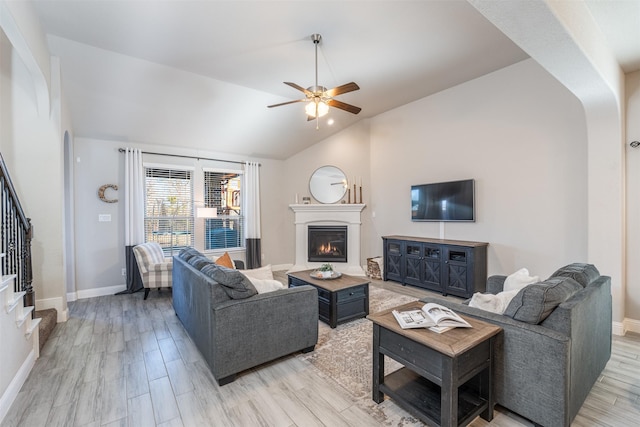 living area with lofted ceiling, a ceiling fan, light wood-type flooring, a warm lit fireplace, and stairs