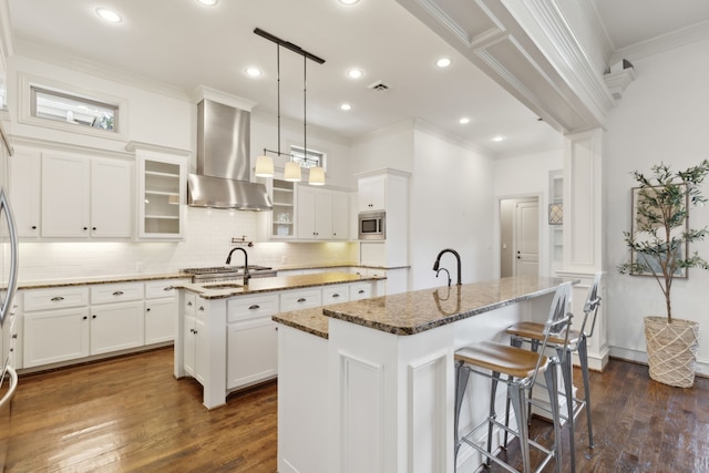 kitchen featuring dark stone counters, a center island with sink, wall chimney range hood, decorative light fixtures, and dark hardwood / wood-style flooring