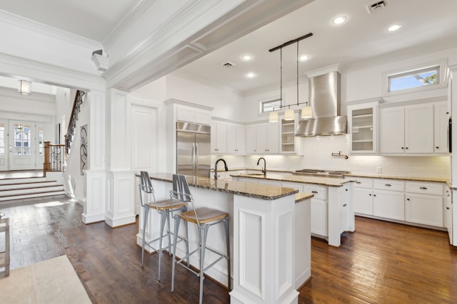 kitchen with dark stone counters, a center island with sink, wall chimney exhaust hood, appliances with stainless steel finishes, and a kitchen bar