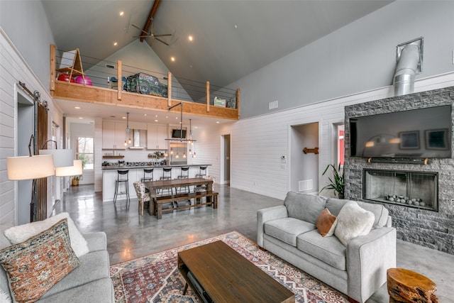 living room featuring high vaulted ceiling, a stone fireplace, ceiling fan, a barn door, and beamed ceiling