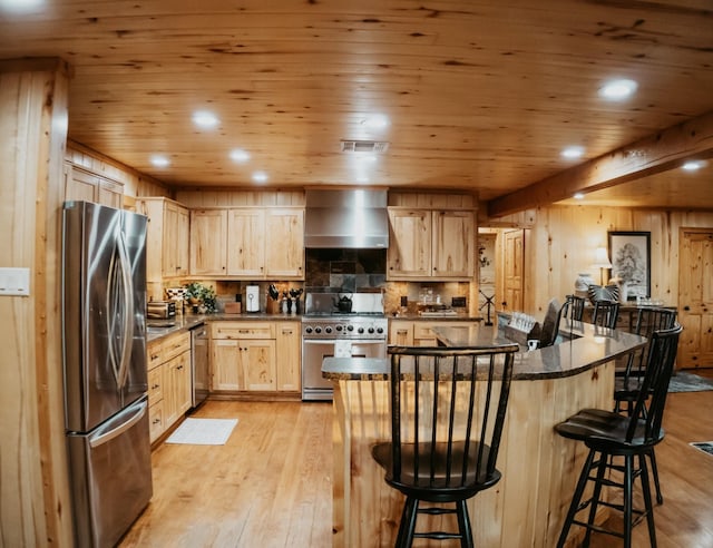 kitchen featuring dark countertops, light wood-style flooring, light brown cabinetry, appliances with stainless steel finishes, and wall chimney range hood