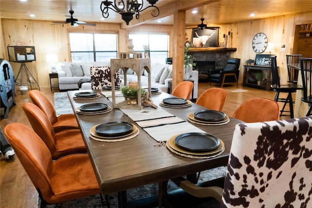 dining space featuring ceiling fan with notable chandelier, wood ceiling, wooden walls, and a stone fireplace