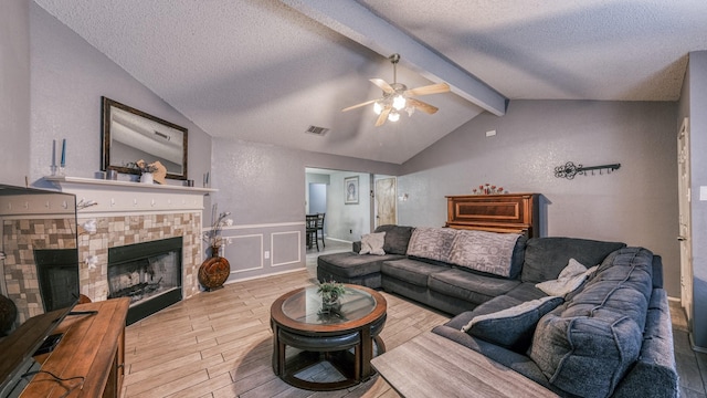 living room with ceiling fan, lofted ceiling with beams, light hardwood / wood-style floors, a textured ceiling, and a tiled fireplace