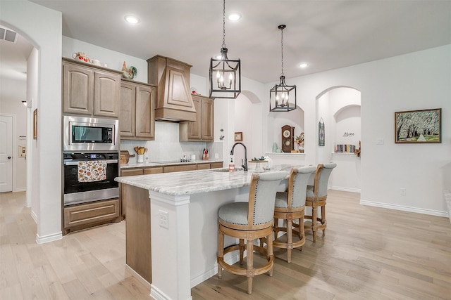 kitchen featuring backsplash, premium range hood, a kitchen island with sink, light stone countertops, and appliances with stainless steel finishes