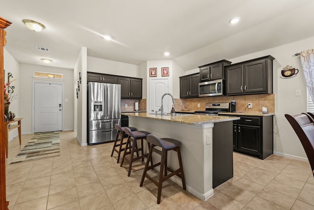 kitchen featuring a kitchen island with sink, sink, light stone countertops, appliances with stainless steel finishes, and tasteful backsplash