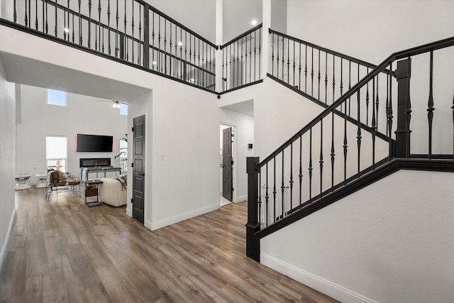 staircase featuring baseboards, a ceiling fan, a glass covered fireplace, a towering ceiling, and wood finished floors