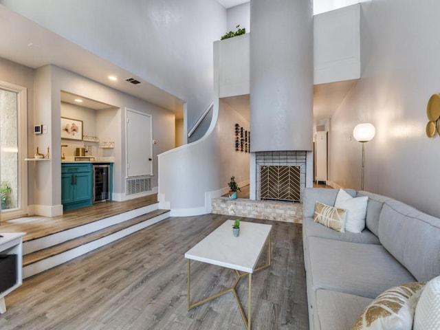 living room with wine cooler, a brick fireplace, light wood-type flooring, and a high ceiling