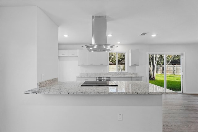 kitchen with island exhaust hood, kitchen peninsula, light stone counters, a wealth of natural light, and white cabinetry